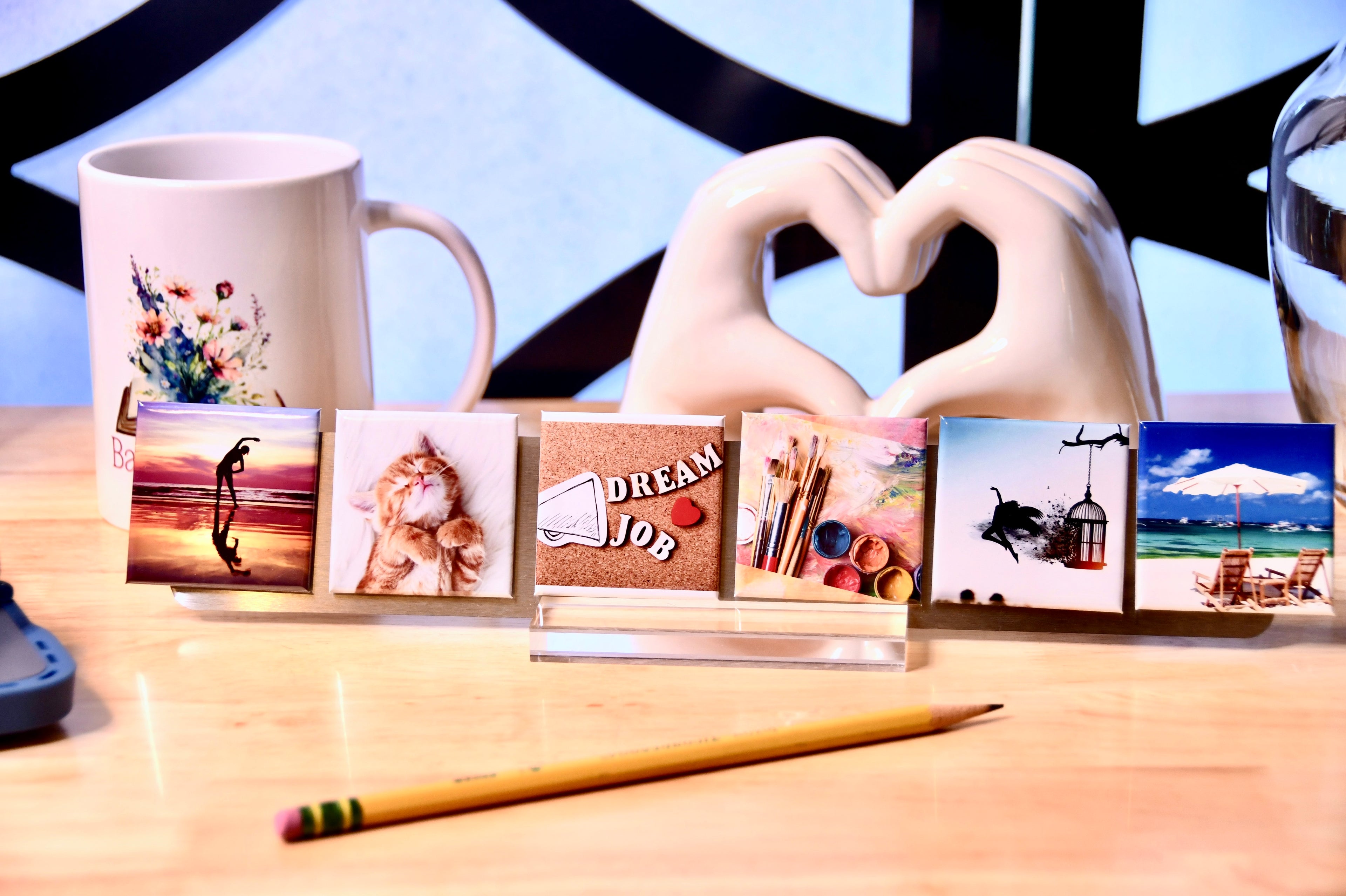A 2" x 12" metal strip seated in a clear acrylic base, holding five 2" x 2" custom photo magnets. A pencil sits in the forefront; a white coffee mug and a ceramic sculpture of heart hands sit in the background.